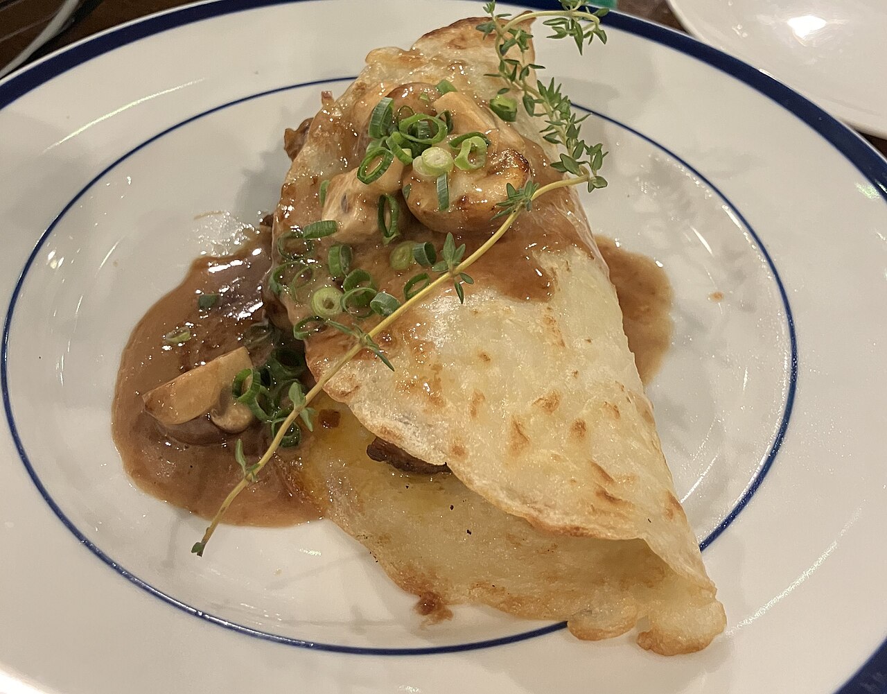 A boxty pancake folded over a mushroom and thyme sauce on a white plate with a blue rim
