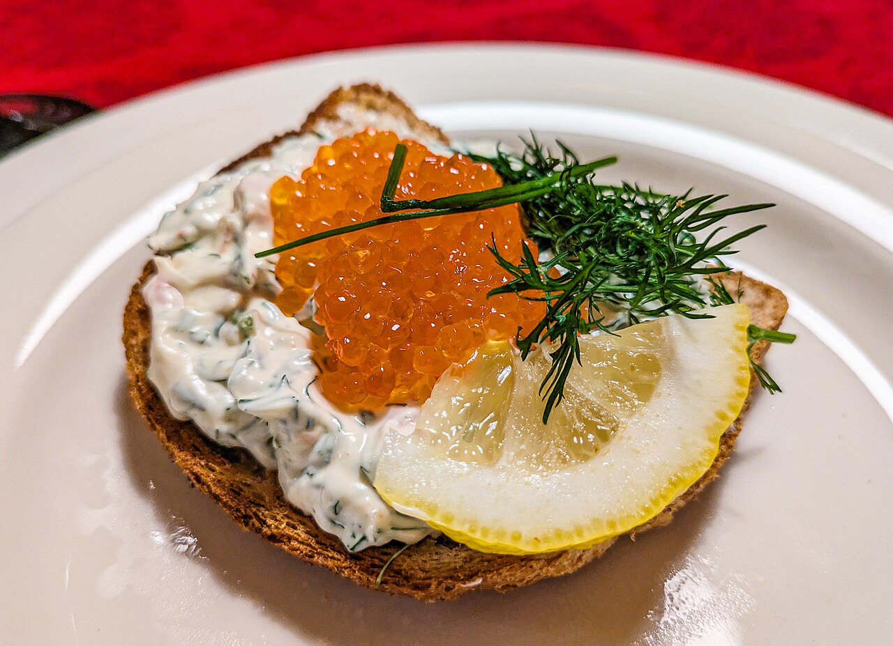 A slice of toast topped with creamy prawn salad, orange roe, dill and a lemon wedge on a white plate