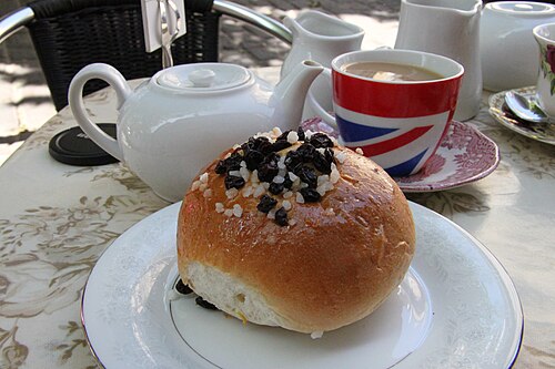 A Bath bun topped with sugar and currants, beside a pot of tea and a cup