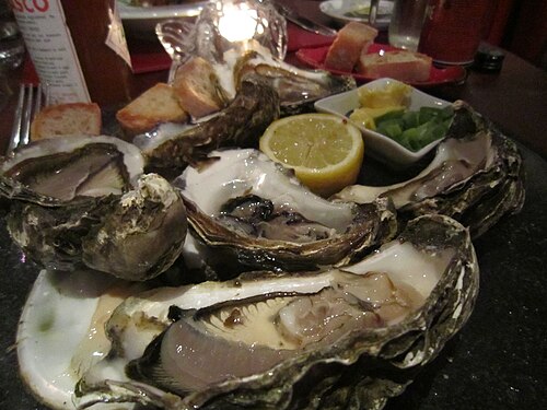 A plate of open oysters on the shell with a lemon wedge and brown bread at the side