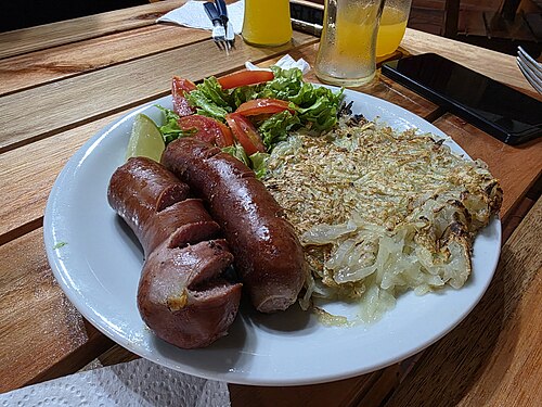 Rösti, a crisp grated-potato cake, on a plate beside two grilled sausages and a salad