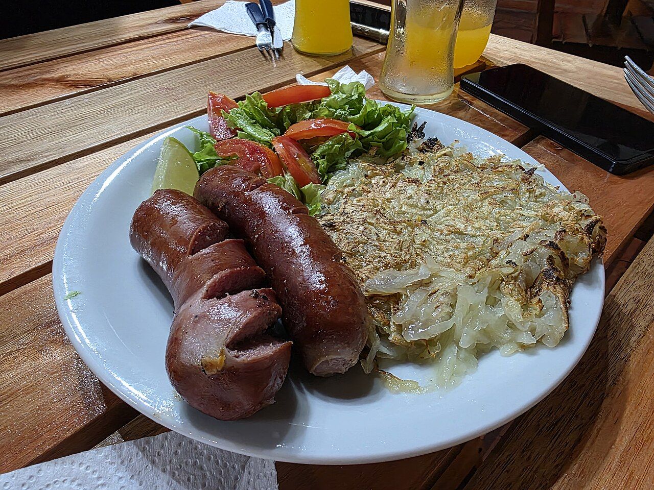 Rösti, a crisp grated-potato cake, on a plate beside two grilled sausages and a salad
