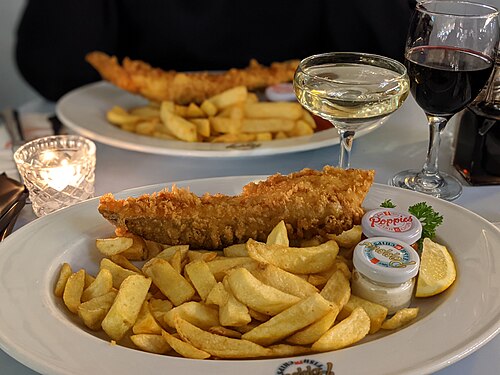 Battered fish and chips on a white plate with a small pot of sauce and a lemon wedge, glasses of wine behind, at a fish and chip restaurant in Spitalfields