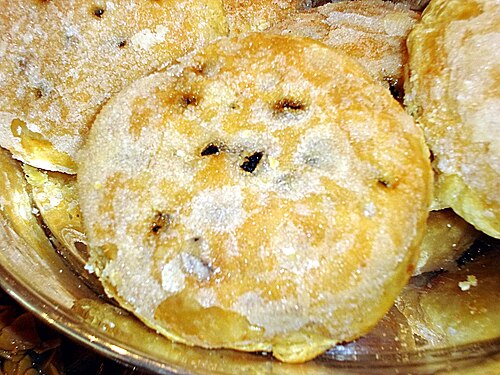 A round Eccles cake with a sugared, flaky pastry top and dried fruit showing through the slits, on a glass dish