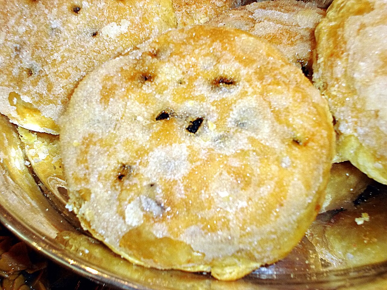 A round Eccles cake with a sugared, flaky pastry top and dried fruit showing through the slits, on a glass dish
