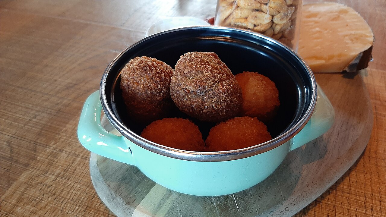 Five bitterballen in a small turquoise pot on a wooden table, with a wedge of cheese behind
