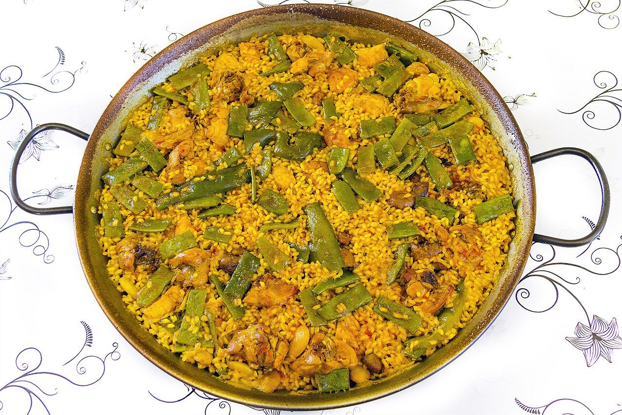 A paella valenciana in its wide pan, saffron rice with chicken pieces and flat green beans, seen from above on a white tablecloth